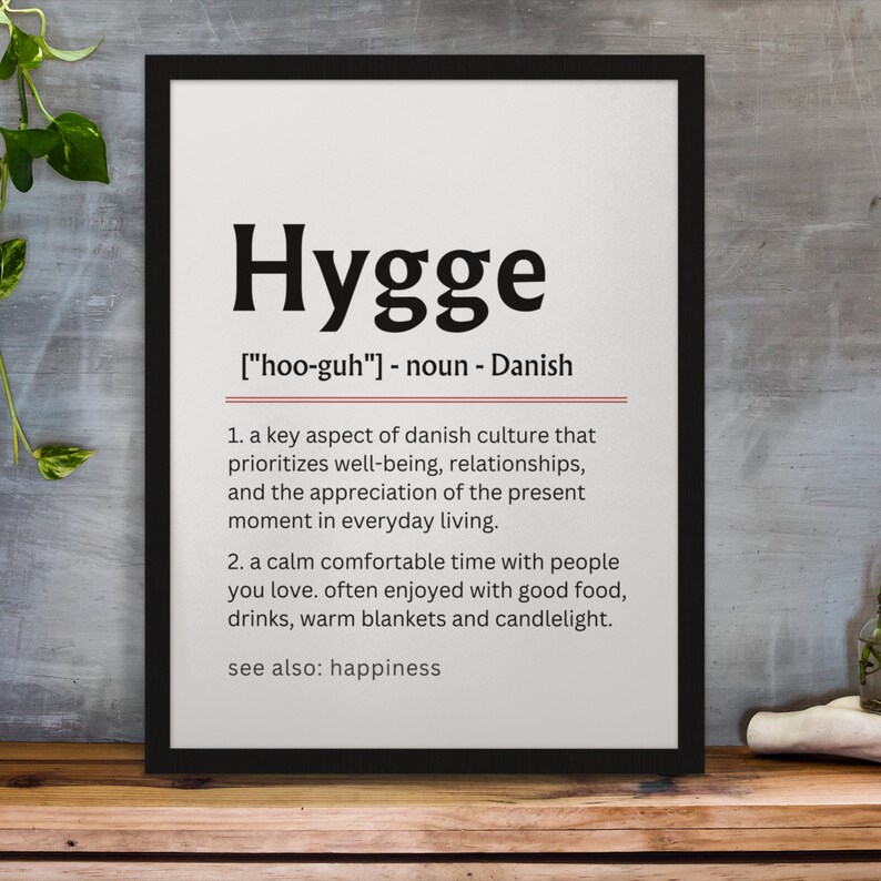 May include: A black framed print with a white background and the word "Hygge" in bold black text. The definition of the word is in smaller black text, with the pronunciation, "[hoo-guh]", and the word's origin, "Danish". The definition is: "A key aspect of Danish culture that prioritizes well-being, relationships, and the appreciation of the present moment in everyday living. A calm comfortable time with people you love. Often enjoyed with good food, drinks, warm blankets and candlelight. See also: happiness."