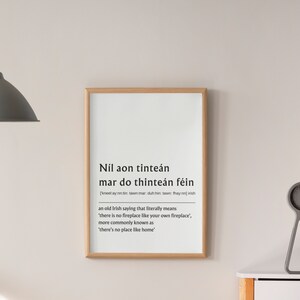 May include: A framed print with a light wood frame. The print features text in both Irish and English. The Irish text is in a bold, sans-serif font. The English translation reads, "there's no place like home."