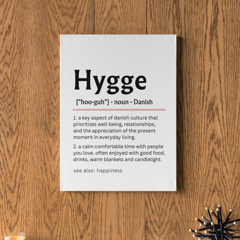 May include: A white canvas print with the word "Hygge" in black bold letters. The definition of the word is written in black text below, including the pronunciation, "[hoo-guh]", and the word's origin, "Danish". The definition is in two parts, with the first part describing Hygge as a key aspect of Danish culture that prioritizes well-being, relationships, and the appreciation of the present moment in everyday living. The second part describes Hygge as a calm, comfortable time with people you love, often enjoyed with good food, drinks, warm blankets, and candlelight. The definition ends with "see also: happiness".