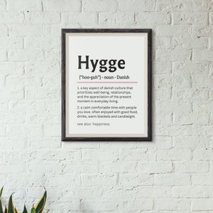 May include: A black framed print with a white background and the word "Hygge" in bold black text. The definition of Hygge is written in black text below the word. The definition includes the pronunciation of the word, "[hoo-guh]", and states that it is a noun of Danish origin. The definition is in two parts, with the first part describing Hygge as a key aspect of Danish culture that prioritizes well-being, relationships, and the appreciation of the present moment in everyday living. The second part describes Hygge as a calm, comfortable time with people you love, often enjoyed with good food, drinks, warm blankets, and candlelight. The definition ends with "see also: happiness."