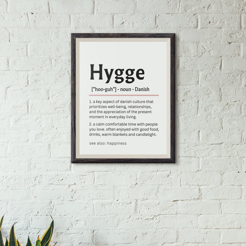 May include: A black framed print with a white background and the word "Hygge" in bold black text. The definition of Hygge is written in black text below the word. The definition includes the pronunciation of the word, "[hoo-guh]", and states that it is a noun of Danish origin. The definition is in two parts, with the first part describing Hygge as a key aspect of Danish culture that prioritizes well-being, relationships, and the appreciation of the present moment in everyday living. The second part describes Hygge as a calm, comfortable time with people you love, often enjoyed with good food, drinks, warm blankets, and candlelight. The definition ends with "see also: happiness."