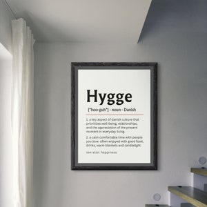 May include: A black framed print with the word "Hygge" in bold white letters. The definition of the word is written in black text below, explaining that it is a Danish word meaning a key aspect of Danish culture that prioritizes well-being, relationships, and the appreciation of the present moment in everyday living. It also describes Hygge as a calm comfortable time with people you love, often enjoyed with good food, drinks, warm blankets and candlelight. The print also states that Hygge is synonymous with happiness.