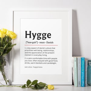May include: A white framed print with the word "Hygge" in black bold letters. The definition of the word is in black text below, with the pronunciation in brackets. The definition explains that Hygge is a key aspect of Danish culture that prioritizes well-being, relationships, and the appreciation of the present moment in everyday living. It also describes Hygge as a calm comfortable time with people you love, often enjoyed with good food, drinks, warm blankets, and candlelight. The print also states that Hygge is synonymous with happiness.