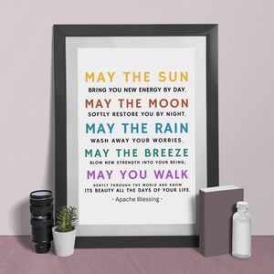 May include: A framed print with the text "May the Sun..." in various colours. The print is in a black frame, with a small plant, a camera lens, a book, and a small bottle next to it. The text is an Apache Blessing.