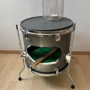 May include: A repurposed drum converted into a side table. The dark gray drum features a black top, chrome hardware, and a cut-out revealing a green interior. A silver wire candle holder sits on top, and drumsticks rest inside.