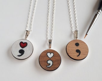 May include: Three silver necklaces with round wooden pendants. The pendants are painted with a white semicolon and a red heart. The necklaces are on a white background.