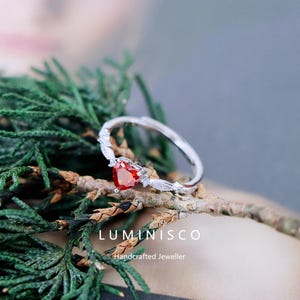 Op de afbeelding: Een zilveren ring met een hartvormige, rode edelsteen. De ring heeft een delicaat, wijnstokachtig ontwerp met kleine, heldere accentstenen. De ring wordt getoond op een natuurlijke tak met groene en bruine bladeren. De tekst "LUMINISCO Handcrafted Jeweller" is zichtbaar.