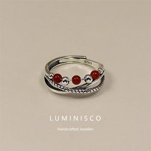 Puede incluir: Anillo ajustable de plata con cinco cuentas rojas y dos cuentas plateadas. El anillo tiene una banda trenzada en forma de cuerda y está estampado con "S925". La marca "LUMINISCO" y "Handcrafted Jeweller" se muestran debajo.