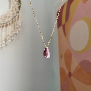 May include: A gold chain necklace with a pendant. The pendant is a teardrop shape with a purple and white colour. The necklace is hanging in front of a wall with a painting and a woven wall hanging.