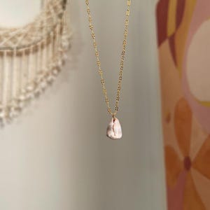 May include: A delicate gold-colored necklace with a small, irregularly shaped, off-white pendant with pinkish hues. The chain has a repeating leaf-like pattern. The necklace hangs against a soft, neutral background, suggesting a focus on minimalist jewelry.