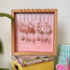 May include: Five gold necklaces with pink seashells and smaller seashells hanging from them. The necklaces are displayed on a wooden frame with a pink and white ombre background.