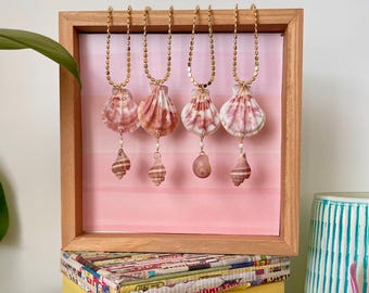 May include: Five gold necklaces with pink seashells and smaller seashells hanging from them. The necklaces are displayed on a wooden frame with a pink and white ombre background.