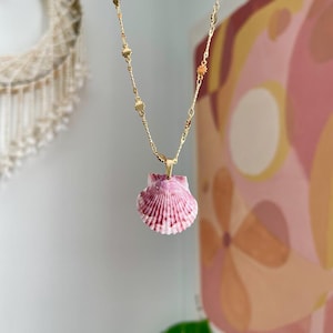 May include: A gold-colored necklace with a pendant made from a pink and white seashell. The chain has small, decorative gold accents. The seashell pendant is a light pink color with darker pink stripes.
