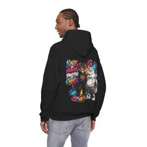 May include: A black hooded sweatshirt featuring a colourful graphic on the back. The design displays two anthropomorphic cats wearing streetwear and the text "KINGS BY NATURE, PAWSITIVE BY CHOICE" surrounded by vibrant paint splatter effects.