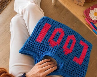 Puede incluir: Un bolso de ganchillo azul con la palabra COOL en letras de bloque de color rosa brillante. El bolso está construido con un patrón de rejilla abierta y tiene asas integradas. El diseño es vibrante y presenta un estilo de fuente pixelada.