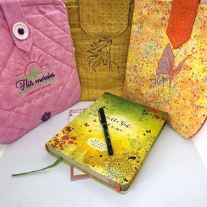 May include: Three quilted fabric pouches in pink, gold, and yellow with embroidered designs, including a horse and a butterfly with the word Hope. A yellow and green journal with the text Hello God it's me and a black pen rest in front.