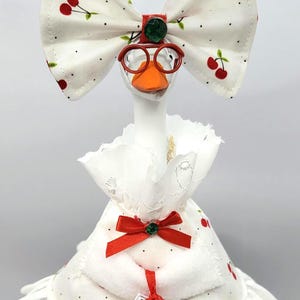 May include: A whimsical goose figurine wearing a white dress with a red bow and cherry print. The goose has orange glasses, a large bow, and a small gift. The figurine is a decorative item.