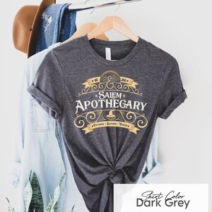 May include: Dark grey t-shirt with rolled-up sleeves. The shirt features a gold and white design that reads "Salem Apothecary" with the text "Potions - Elixirs - Tonics". The shirt is hanging on a wooden hanger.