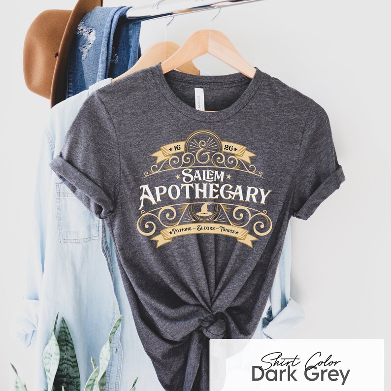 May include: Dark grey t-shirt with rolled-up sleeves. The shirt features a gold and white design that reads "Salem Apothecary" with the text "Potions - Elixirs - Tonics". The shirt is hanging on a wooden hanger.