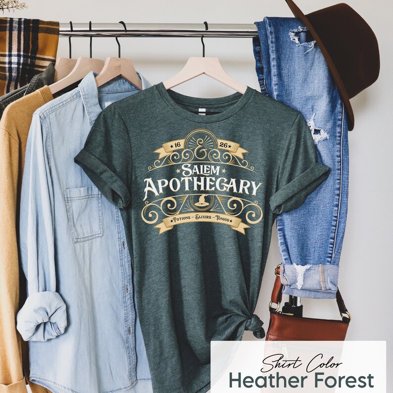 May include: A heather forest green t-shirt with the text "Salem Apothecary" in a decorative design. The shirt has rolled-up sleeves and is hanging on a clothing rack with other garments. The text includes "Potions - Elixirs - Tonics".