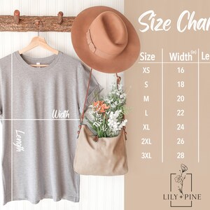 May include: A light gray t-shirt hangs on a wooden hanger, with a size chart to the right. The chart lists sizes XS to 3XL, with width and length measurements in inches. A brown hat and purse with flowers are also visible.