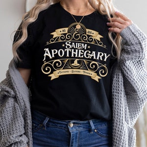 May include: Black t-shirt with a gold and white design that reads "Salem Apothecary". The design includes a witch's hat, stars, and the words "Potions - Elixirs - Tonics". The shirt is a crew neck.