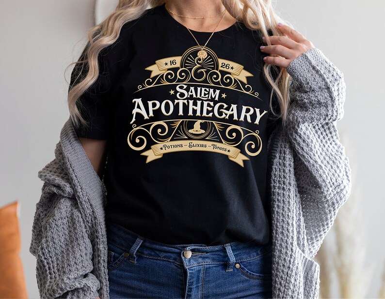May include: Black t-shirt with a gold and white design that reads "Salem Apothecary". The design includes a witch's hat, stars, and the words "Potions - Elixirs - Tonics". The shirt is a crew neck.