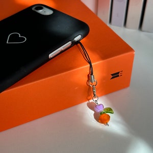 May include: Black phone case with a white heart design. A decorative charm with purple, green, and orange beads hangs from the phone. The phone case rests on an orange box.