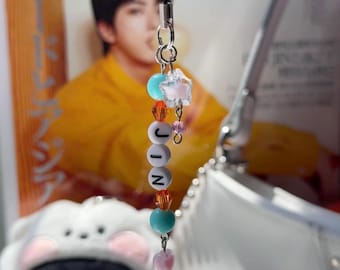 May include: A beaded charm spelling "JIN" in white letter beads. The charm includes a teal bead, a star-shaped bead, and orange beads. It is suspended from a silver-coloured metal loop and a black cord.