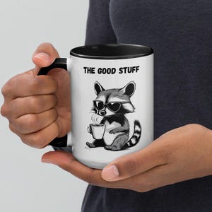 Op de afbeelding: Een witte keramische koffiemok met een zwarte rand en handvat. De mok heeft een zwart-witte illustratie van een wasbeer met een zonnebril die een kopje koffie vasthoudt. De tekst "THE GOOD STUFF" is in zwart op de mok gedrukt.