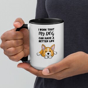 Può includere: Una tazza in ceramica bianca con bordo e manico neri. La tazza presenta un corgi di cartone animato e il testo "Lavoro perché il mio cane possa avere una vita migliore."
