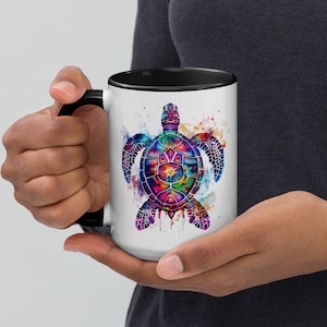 Puede incluir: Una taza de café de cerámica blanca con un borde y un asa negros. La taza presenta una ilustración acuarela colorida de una tortuga marina con un diseño de mandala.