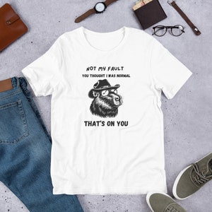 Könnte beinhalten: Weißes T-Shirt mit einer schwarz-weißen Illustration eines Bären, der einen Cowboyhut und eine Sonnenbrille trägt. Der Text "Not my fault you thought I was normal. That's on you" ist auf dem T-Shirt aufgedruckt.