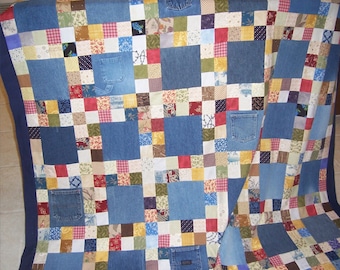 May include: A patchwork quilt made from denim fabric with various colours and patterns. The quilt is made up of squares of different sizes and colours, including blue, red, white, and brown. The quilt is folded in half and is lying on a table.