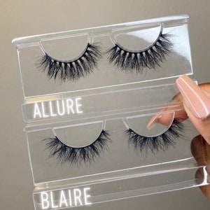 Op de afbeelding: Twee paar valse wimpers in een doorzichtige plastic verpakking. Het bovenste paar is gelabeld "ALLURE" en het onderste paar is gelabeld "BLAIRE".