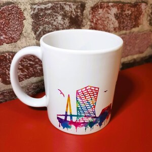 Peut inclure: Mug en céramique blanche avec un motif de paysage urbain coloré, représentant une silhouette de bâtiment et de pont. Le motif comprend des gouttes vibrantes de style aquarelle dans les tons de bleu, rose, jaune et violet. Deux oiseaux volent dans le ciel.