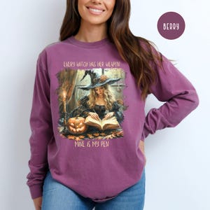 May include: Berry-colored long-sleeve shirt featuring a witch illustration. The graphic shows a witch reading a book with a pumpkin and the text "EVERY WITCH HAS HER WEAPON, MINE IS MY PEN". The shirt is a comfortable, casual style.