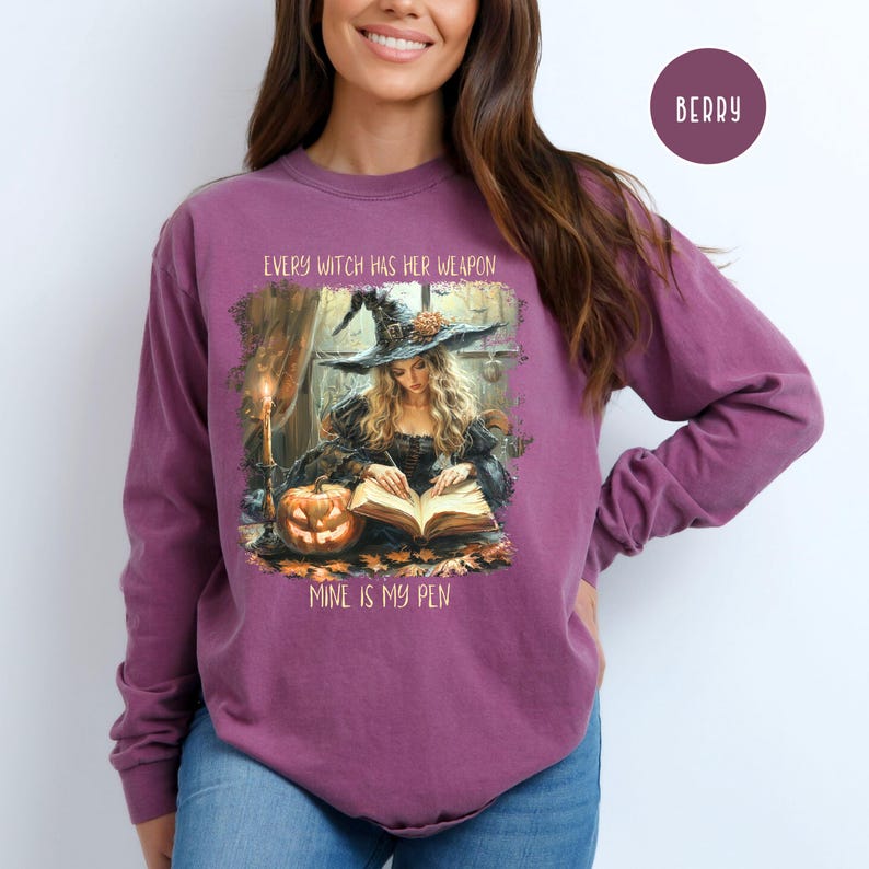 May include: Berry-colored long-sleeve shirt featuring a witch illustration. The graphic shows a witch reading a book with a pumpkin and the text "EVERY WITCH HAS HER WEAPON, MINE IS MY PEN". The shirt is a comfortable, casual style.