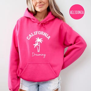 Puede incluir: Una sudadera con capucha de color rosa brillante con un estampado gr&aacute;fico blanco que dice "CALIFORNIA DREAMING" con palmeras.