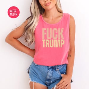 K&ouml;nnte beinhalten: Ein pinkfarbenes Tanktop mit den Worten "FUCK TRUMP" in Beige aufgedruckt. Das Tanktop wird mit Jeansshorts kombiniert. Ein "WATER-MELON"-Aufkleber befindet sich in der oberen linken Ecke.