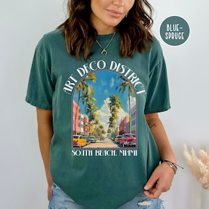 May include: A teal blue t-shirt with a graphic print of a street scene in Miami's Art Deco District. The text "Art Deco District" is printed in a white arch above the image. The text "South Beach, Miami" is printed below the image.