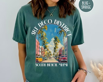 May include: A teal blue t-shirt with a graphic print of a street scene in Miami's Art Deco District. The text "Art Deco District" is printed in a white arch above the image. The text "South Beach, Miami" is printed below the image.