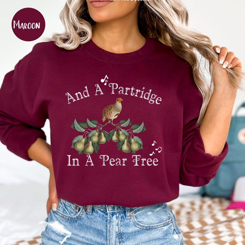 May include: A maroon sweatshirt with the text "And A Partridge In A Pear Tree" in white. A small illustration of a partridge on a pear tree branch with pears is centered on the sweatshirt.