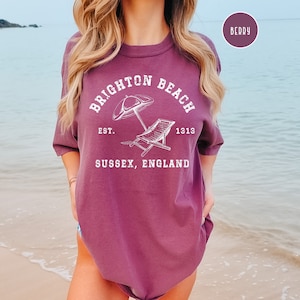 K&ouml;nnte beinhalten: Ein T-Shirt in Beerent&ouml;nen mit einem wei&szlig;en Grafikdesign, das den Text "Brighton Beach Est. 1913 Sussex, England" sowie einen Sonnenschirm und einen Liegestuhl zeigt.