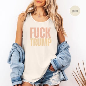 K&ouml;nnte beinhalten: Elfenbeinfarbenes Tanktop mit dem Text "FUCK TRUMP" in rosa und gelben Buchstaben. Das Tanktop wird mit einer Jeansjacke und Jeansshorts getragen. Das Tanktop ist ein l&auml;ssiges Kleidungsst&uuml;ck.