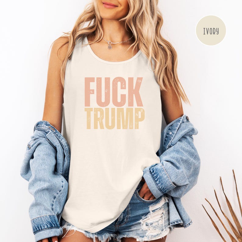 K&ouml;nnte beinhalten: Elfenbeinfarbenes Tanktop mit dem Text "FUCK TRUMP" in rosa und gelben Buchstaben. Das Tanktop wird mit einer Jeansjacke und Jeansshorts getragen. Das Tanktop ist ein l&auml;ssiges Kleidungsst&uuml;ck.