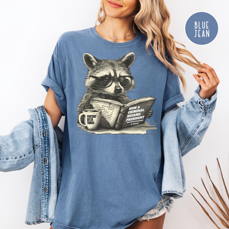 May include: Blue jean-colored t-shirt featuring a raccoon wearing glasses and reading a book titled "How a Criminal Became President." A coffee mug with the text "Foxtrot Delta Tango" sits beside the book. The shirt has rolled-up sleeves.