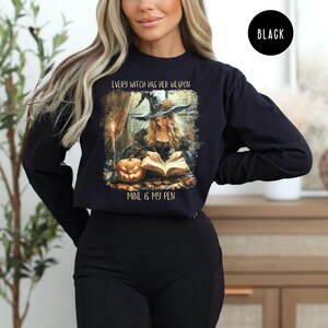 May include: Black long-sleeve sweatshirt featuring a witch illustration with the text "EVERY WITCH HAS HER WEAPON MINE IS MY PEN". The design includes a witch writing in a book, a pumpkin, and a candle.