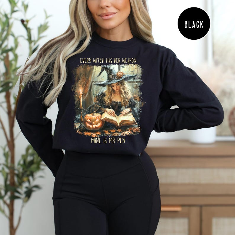 May include: Black long-sleeve sweatshirt featuring a witch illustration with the text "EVERY WITCH HAS HER WEAPON MINE IS MY PEN". The design includes a witch writing in a book, a pumpkin, and a candle.