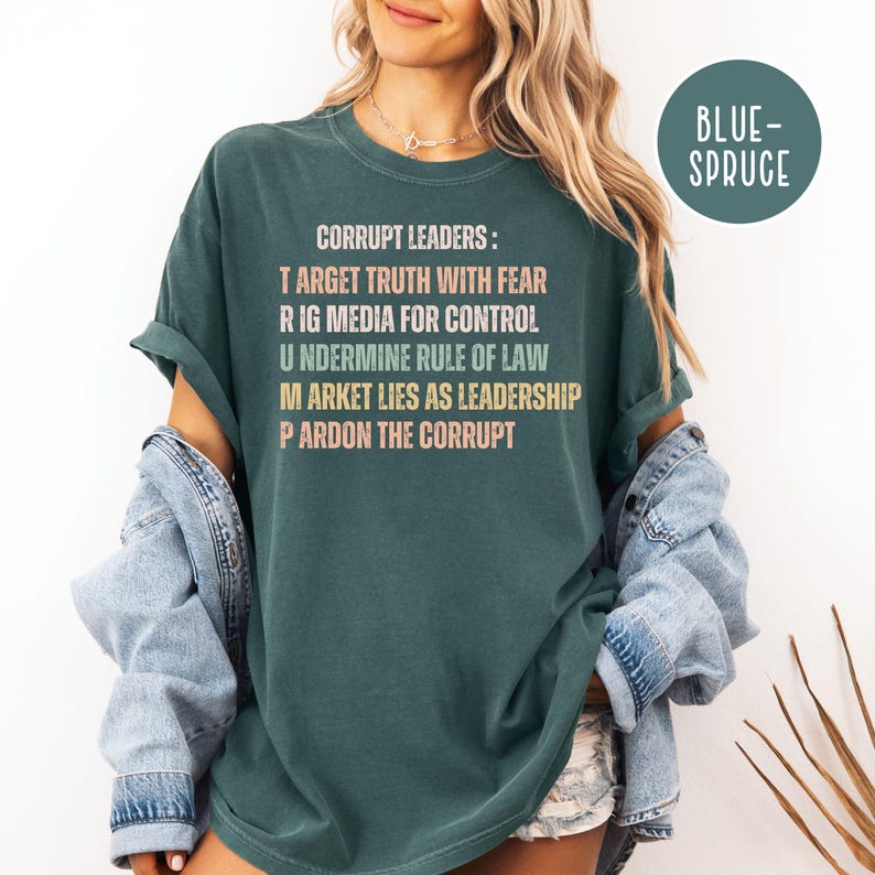May include: Dark teal t-shirt with the text "CORRUPT LEADERS: TARGET TRUTH WITH FEAR, RIG MEDIA FOR CONTROL, UNDERMINE RULE OF LAW, MARKET LIES AS LEADERSHIP, PARDON THE CORRUPT." The words are in various colors. A blue-spruce circle is in the upper right.